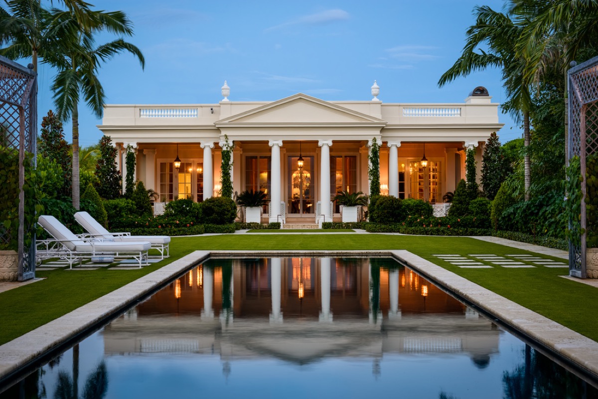 Neoclassical Palm Beach villa at dusk, a columned portico mirrored in a long reflecting pool framed by palms